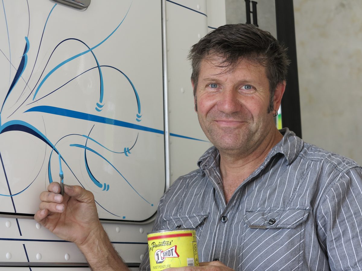 Tony Gibbs holding a brush and a tin of paint beside blue pinstripe scroll work